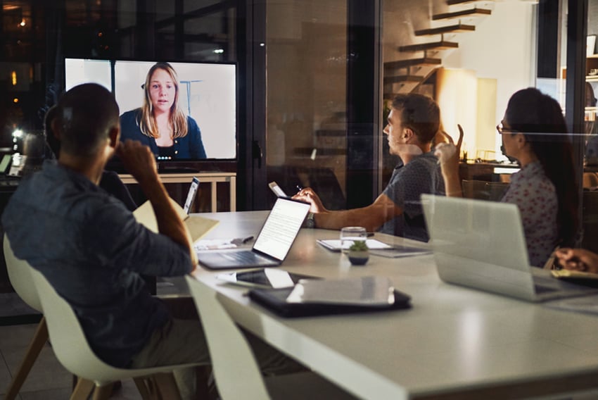 professional men and women at a conference table in an office setting looking at a remote female colleague on a video screen 